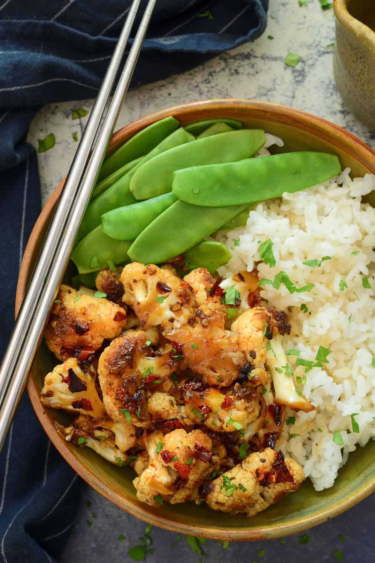 A brown-green ceramic bowl filled with cauliflower stir fry, rice and snow peas. Metal chopsticks rest on the side of the bowl.