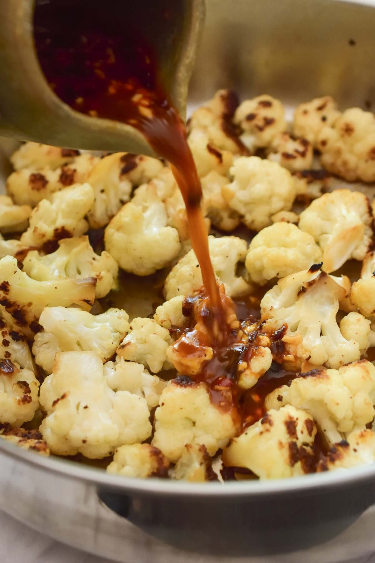 Pouring stir fry sauce into the silver pan of charred cauliflower florets.