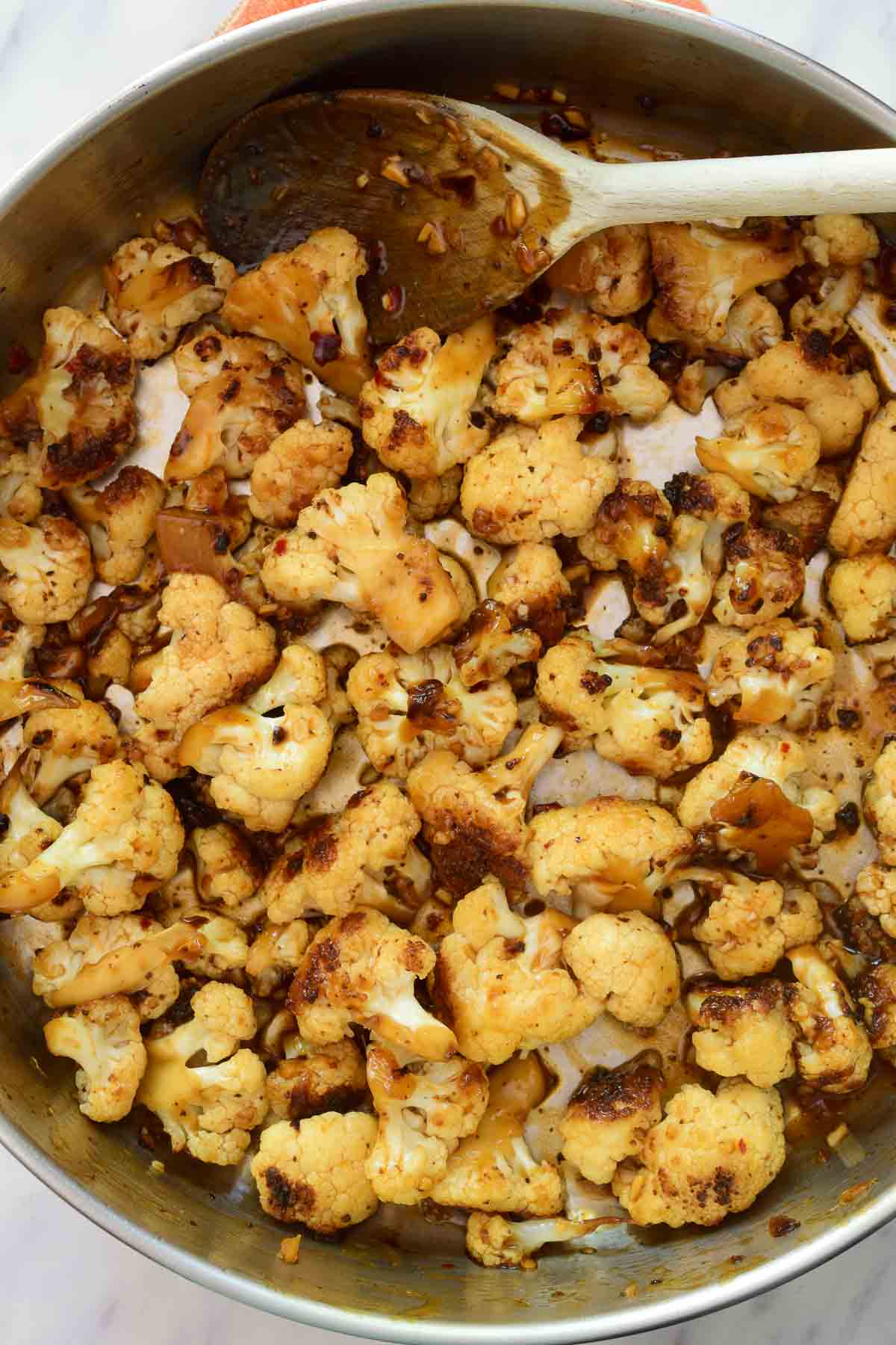 A pan filled with cauliflower stir fry. A wooden spoon rests at the edge of the pan.