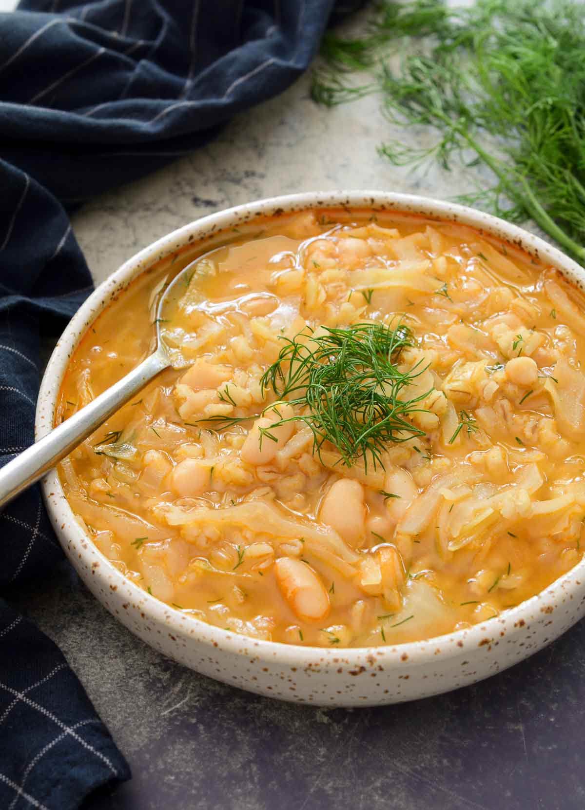 A white ceramic bowl filled with cabbage and barley soup with a pile of dill in the middle and a spoon resting on the side of the bowl.