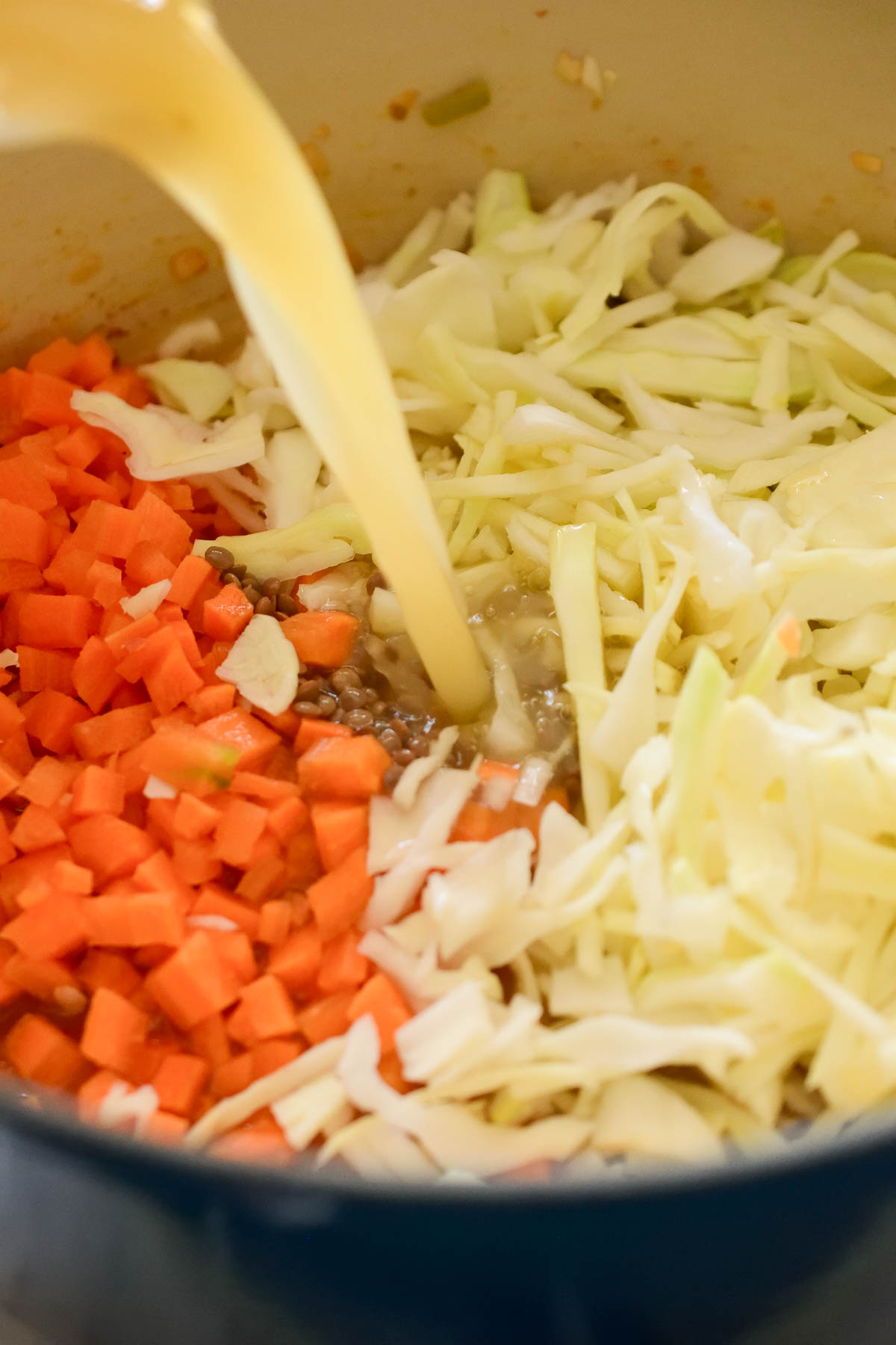 A stream of vegetable stock being poured into a blue pot with diced carrot and shredded cabbage inside.