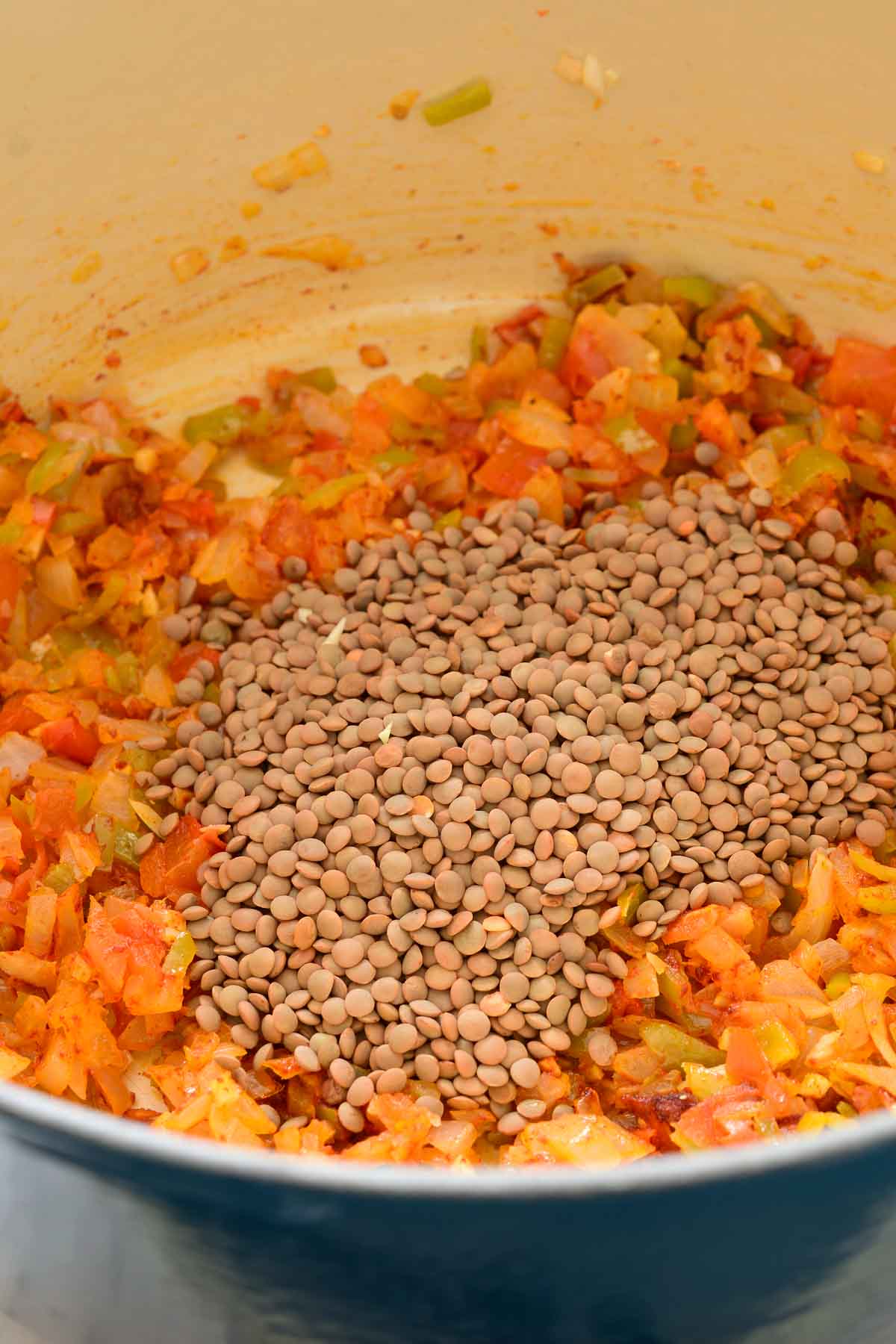A pile of dried brown lentils in the blue pot on top of the fried onion, green pepper and tomato.