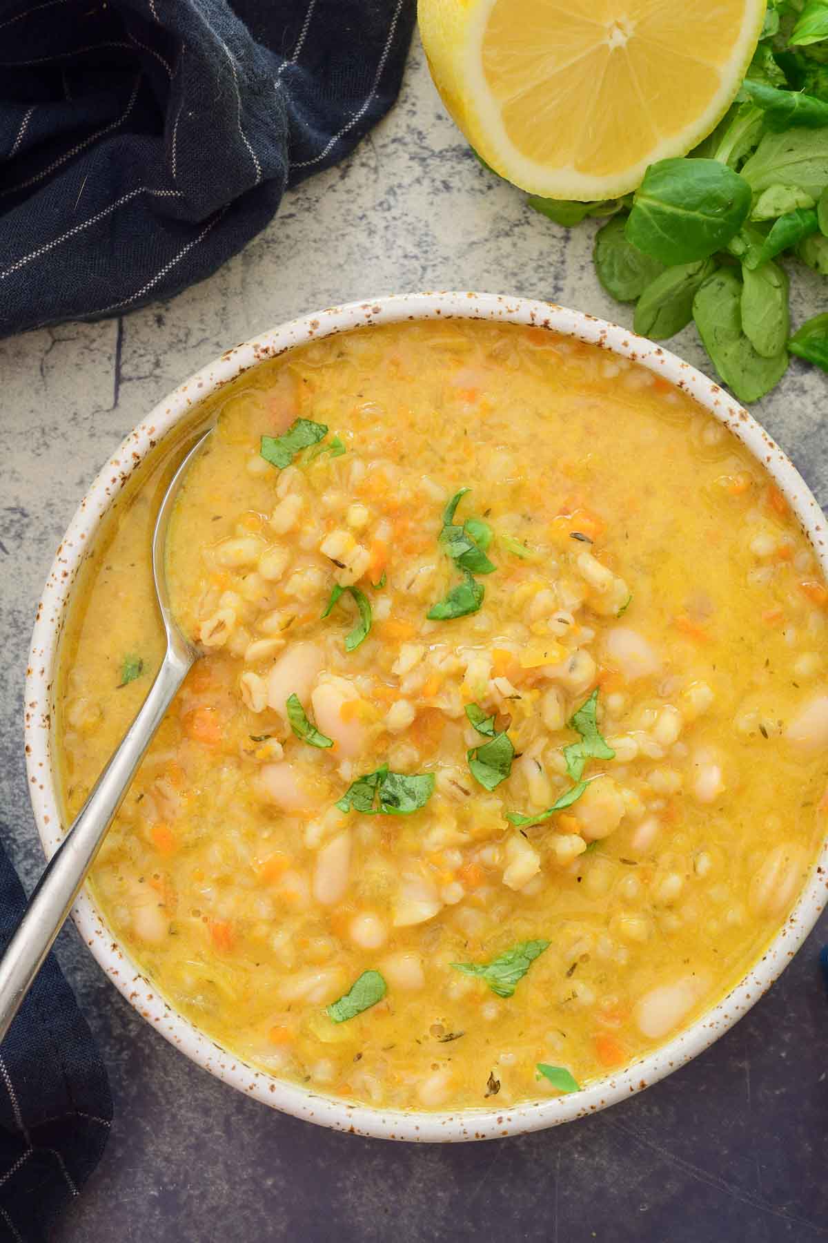 A bowl of lemon barley soup with a spoon in it in a spotted white ceramic bowl. Some greens and a half of lemon surround the bowl.