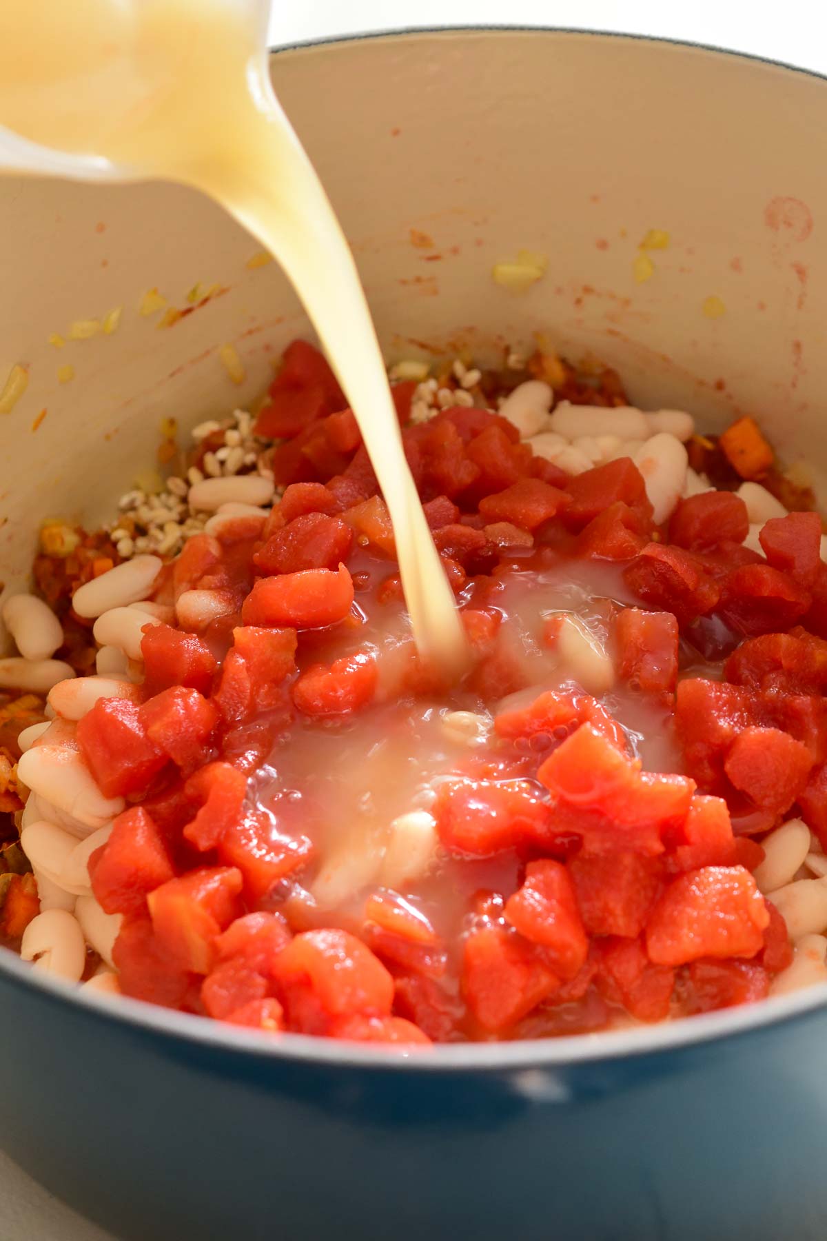 Pouring vegetable stock into a pot with diced tomatoes, beans and barley.