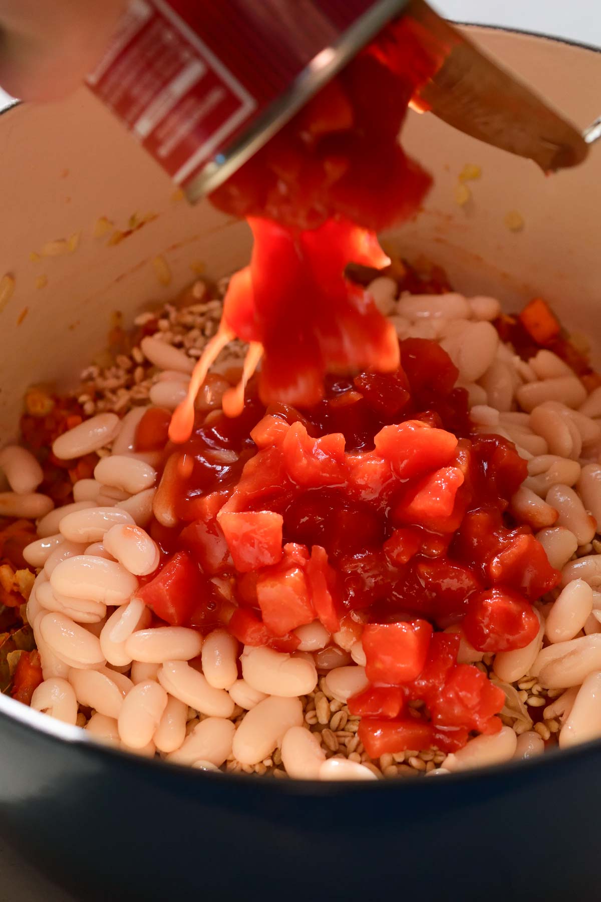 Pouring a can of diced tomatoes into a blue pot of beans, barley and fried vegetables.