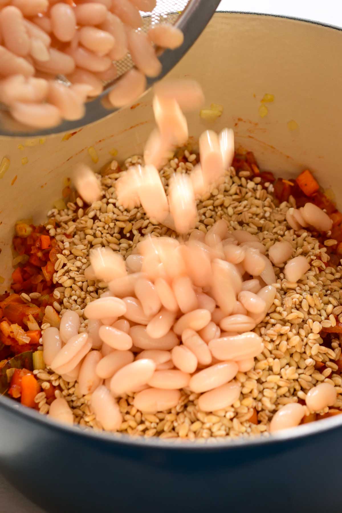 Pouring white beans into a blue pot with barley and fried vegetables.