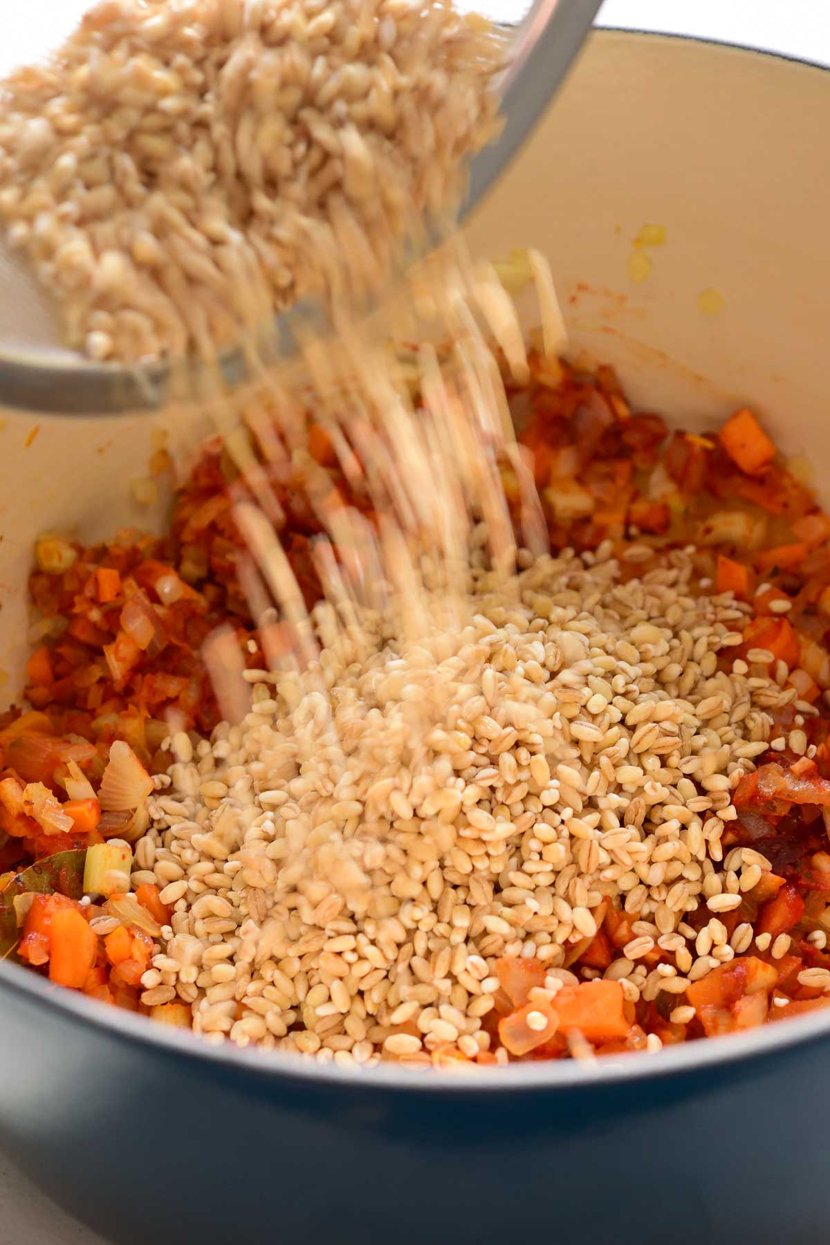 Pouring dried barley into a blue pot with the fried vegetables.