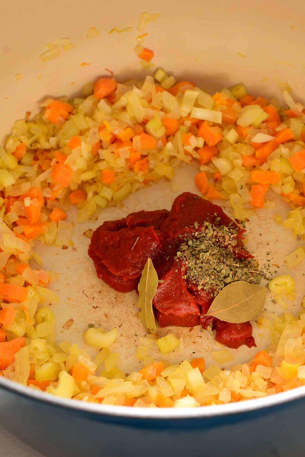 A blue pot filled with fried onion, carrot and celery. Dollops of tomato paste with dried basil and bay leaves in the middle of the pot.